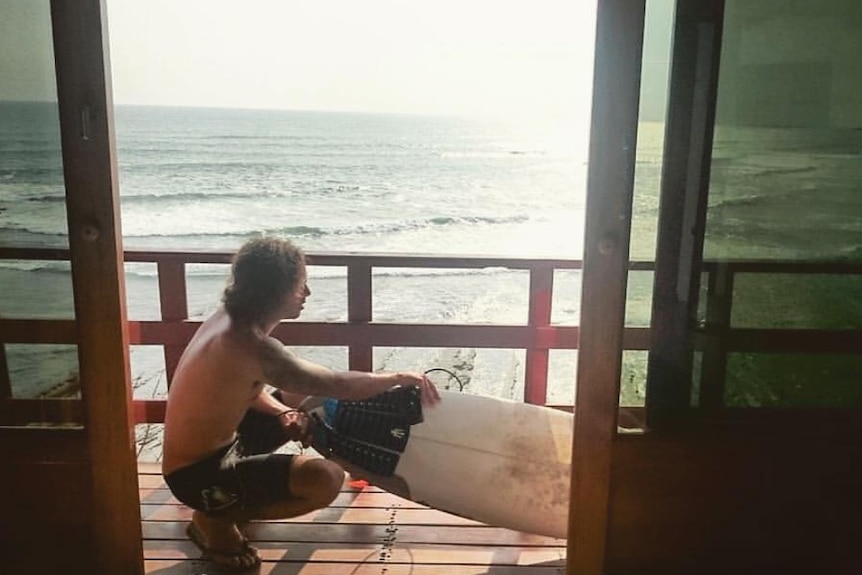 A man crouching and touching a surfboard.