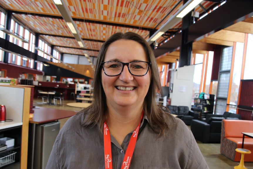 A woman wearing a grey button up and an orange lanyard stand in a classroom, smiling.