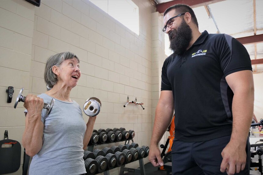 Woman with grey hair holding dumbbells standing next to tall man with beard in a gym.
