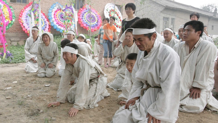 People in white kneel in mourning. There are houses and wreaths in the background.