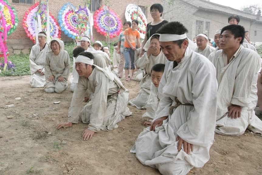 People in white kneel in mourning. There are houses and wreaths in the background.