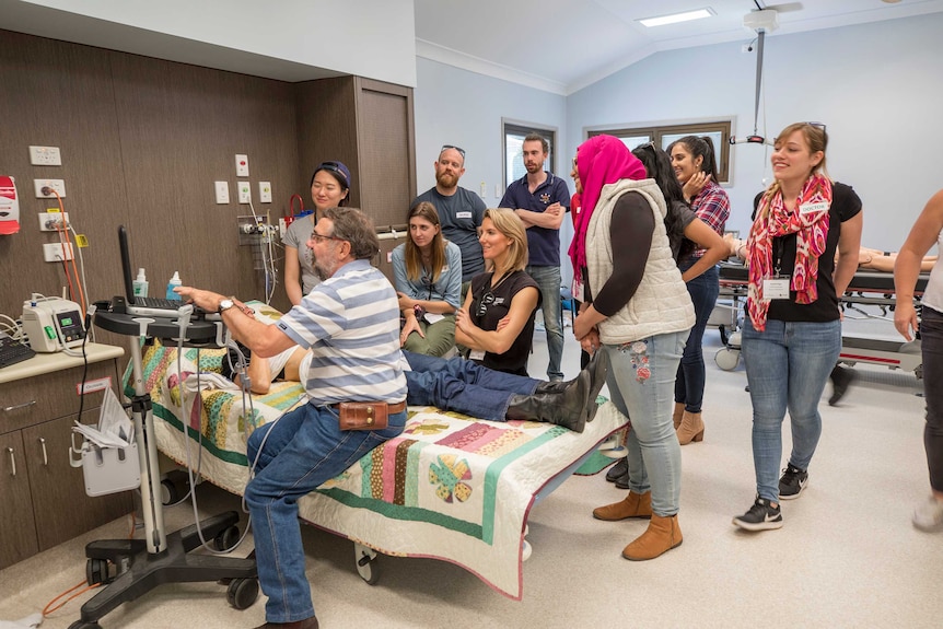 A group of students watch on as a doctor shows them how to use an ultrasound machine on a pretend patient.