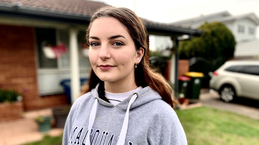 Woman in Macquarie University hoodie looks at the camera outside a house.