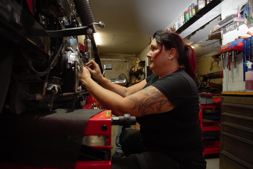 Mechanic Sharine Milne tinkers on motorbike in her workshop, large tattoo on her arm.