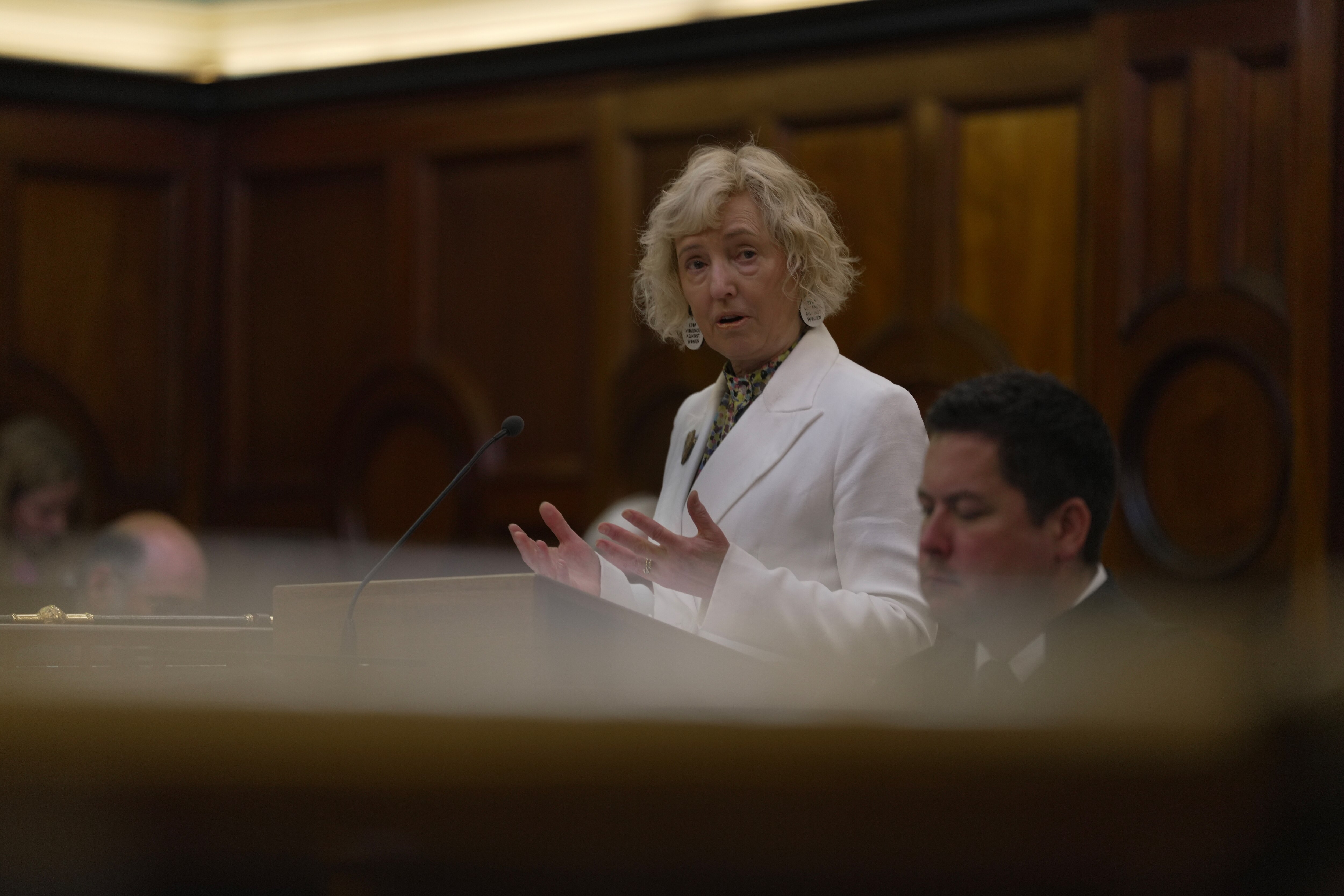 A blonde woman with a white jacket speaking at a lectern.