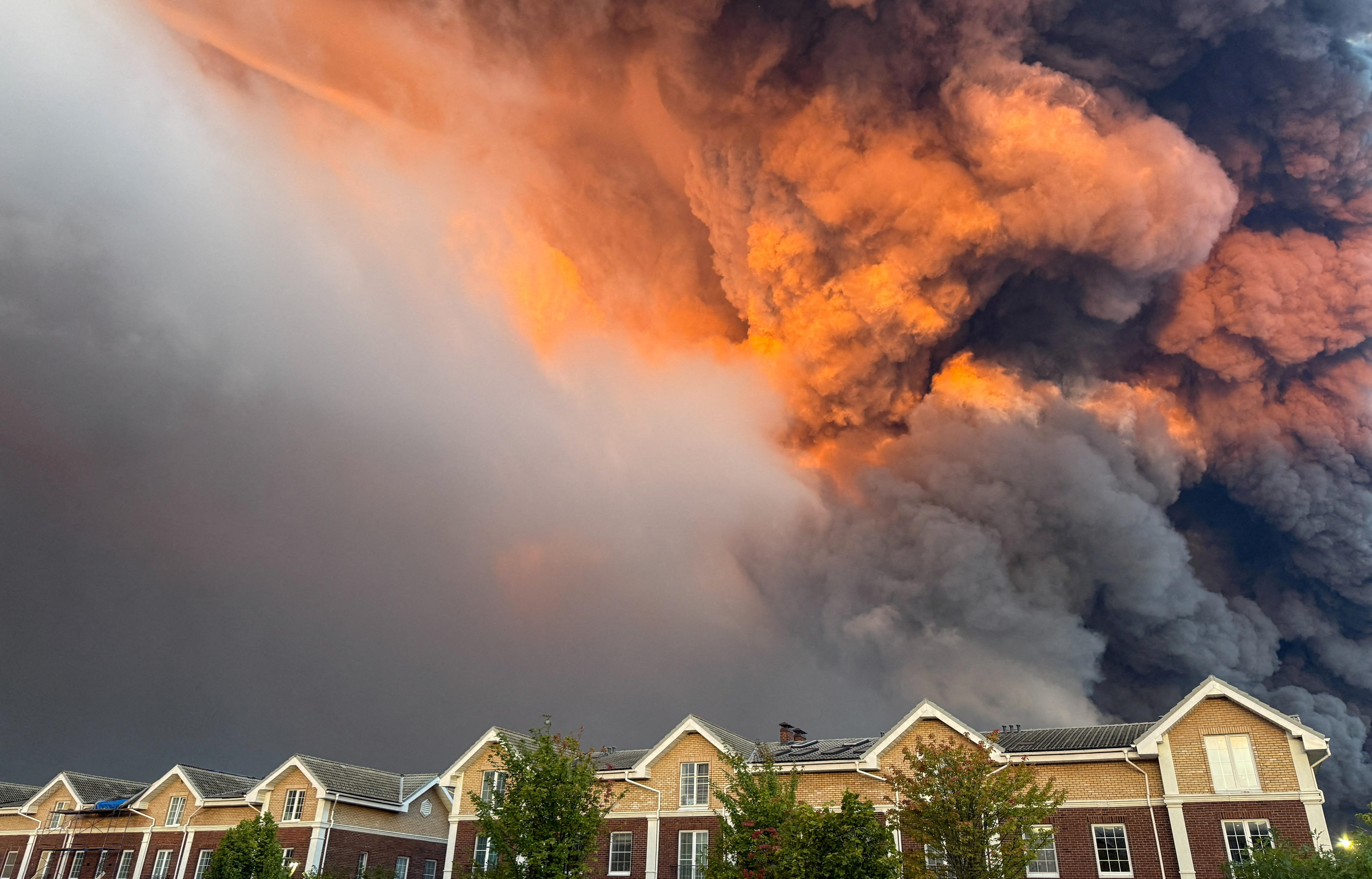 A large plume of smoke reaching high into the atmosphere is seen behind a row of two-storey suburban homes.