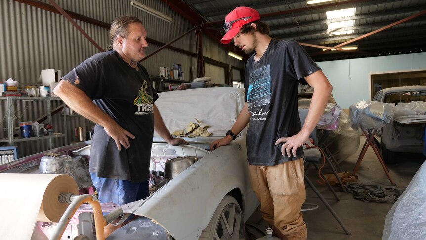 Two men stand over the bonnet of a car in a garage.