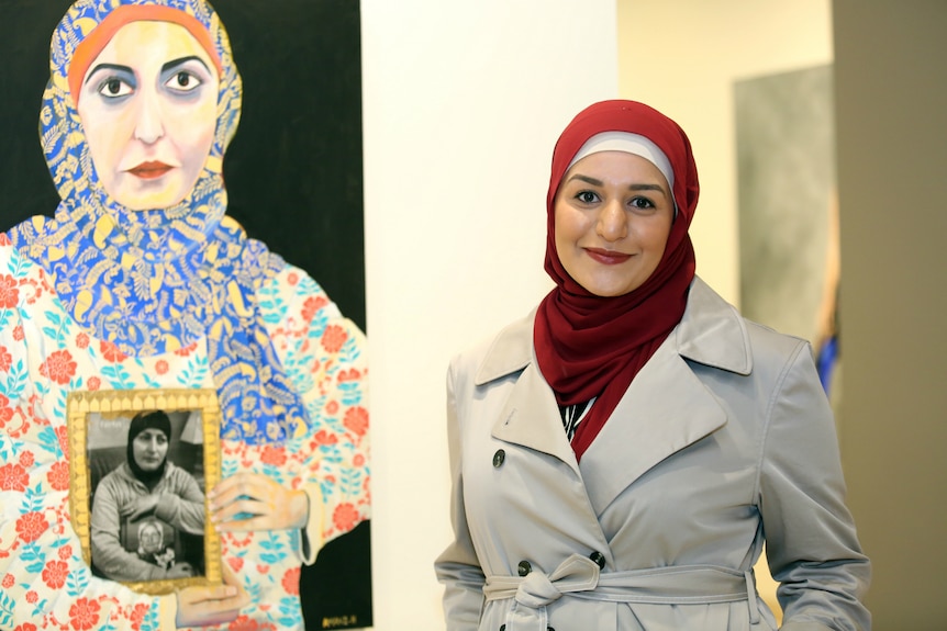 A photo of Amani Haydar in a red hijab, standing in front of her painted self-portrait