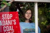Jean Hinchliffe inside her house seen through a window, she is holding a sign calling for the halting of the Adani development.