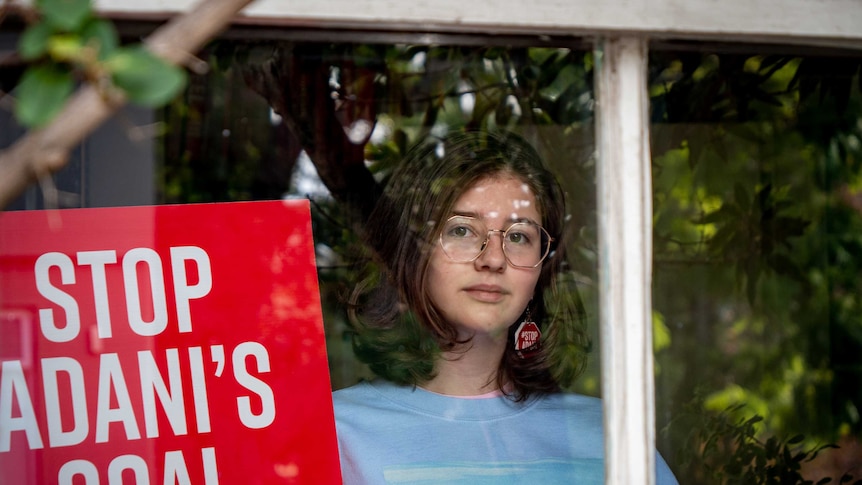 Jean Hinchliffe inside her house seen through a window, she is holding a sign calling for the halting of the Adani development.