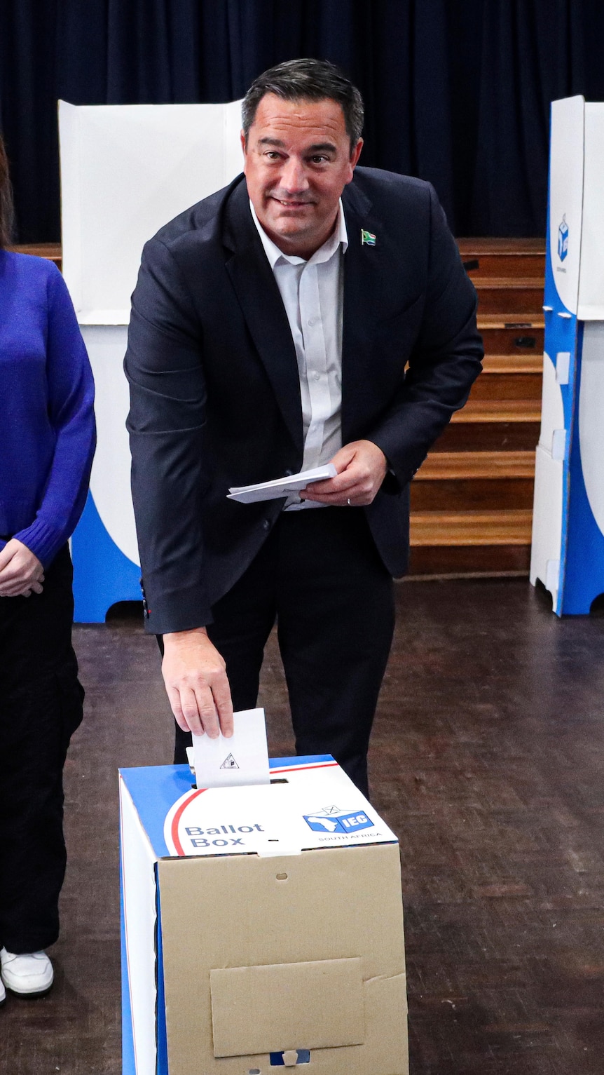 A middle-aged white South African man in a suit smiles as he leans down to place a folded piece of paper in a ballot box.