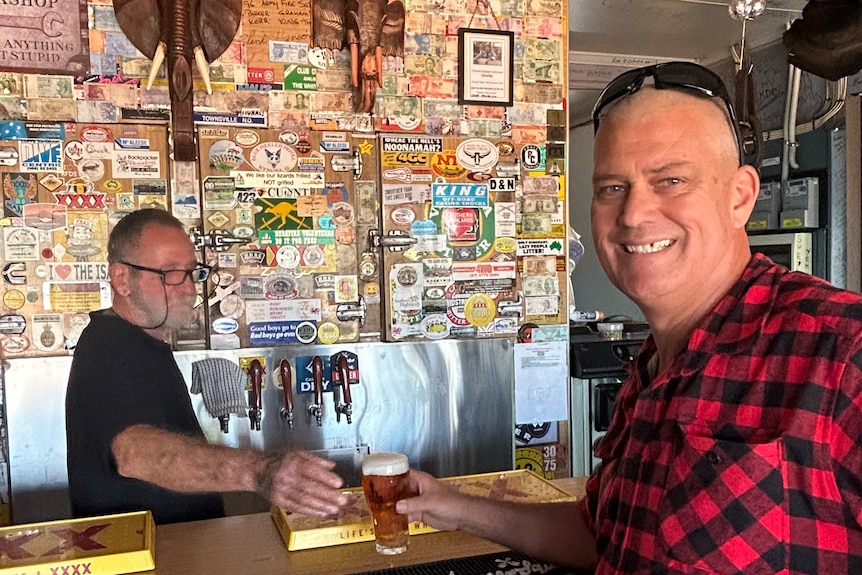 Man in red and black checked shirt stands at bar smiling as man behind the bar hands him a beer.