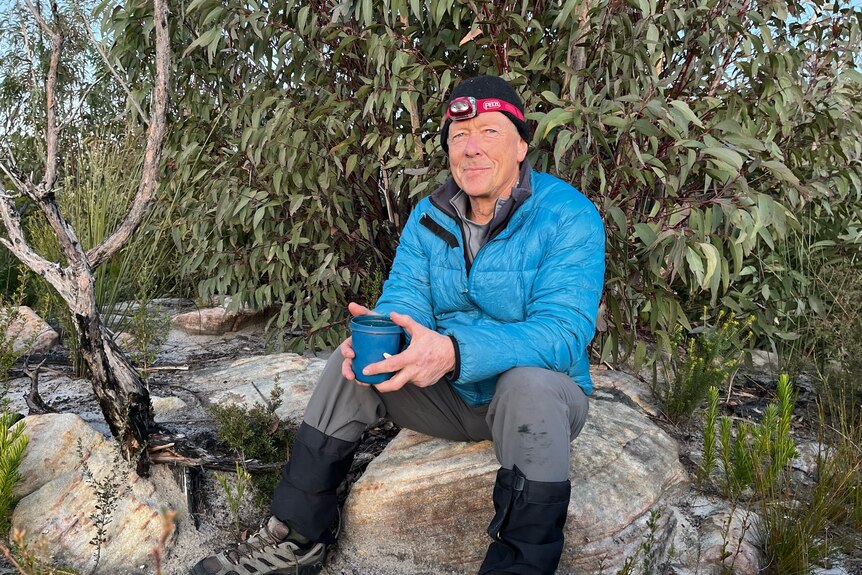 A man with a blue puffer jacket, beanie and head torch sits on a log with a tree behind him.