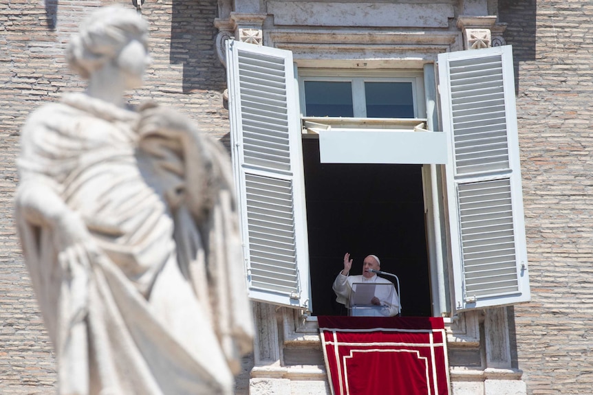 The Pope stands at a high window and gestures as he speaks.