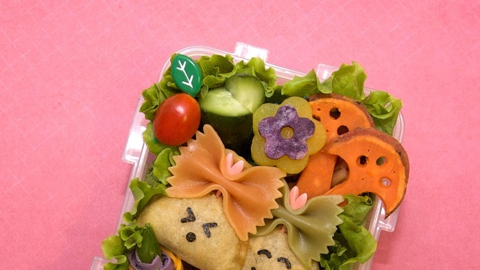 Two pastries with seaweed faces sitting in a lunchbox with vegetables cut into shapes.