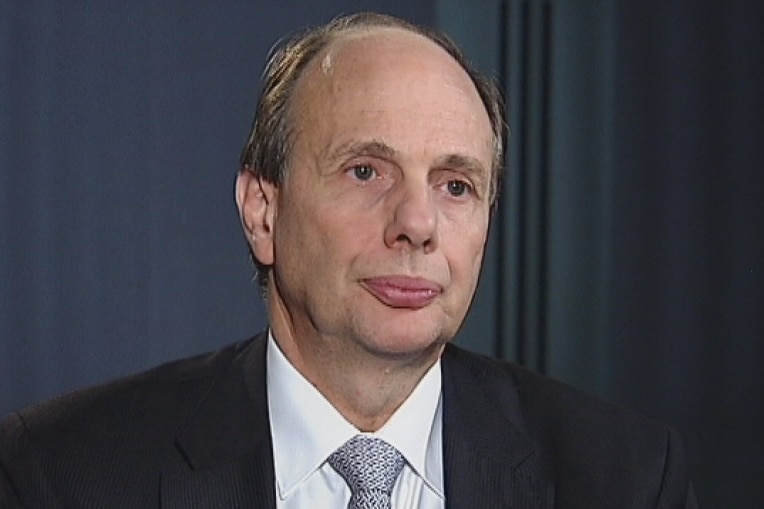 A man in a black suit, white shirt, tie sits in front of a dark background. 