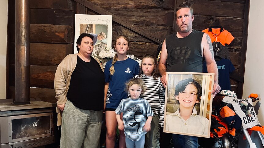A family of five - a man, woman and three young girls hold the portrait of their only son and brother against an orange memorial