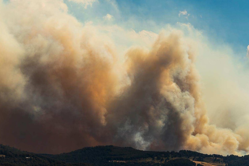 Orangey smoke billows from behind mountains into the blue sky.