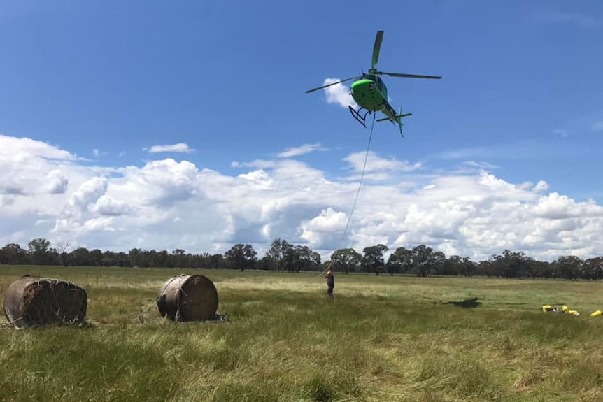 A helicopter with a line trailing from it hovers over hay bales in a field.