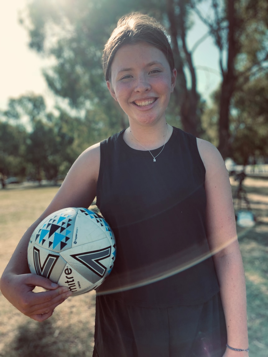 Teenager Harriet pictured outdoors in a leafy park, smiling at the camera and holding a soccer ball under her arm.