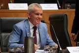 Malcolm Roberts wears a light blue suit and smiles while holding a pen in his right hand during senate estimates.