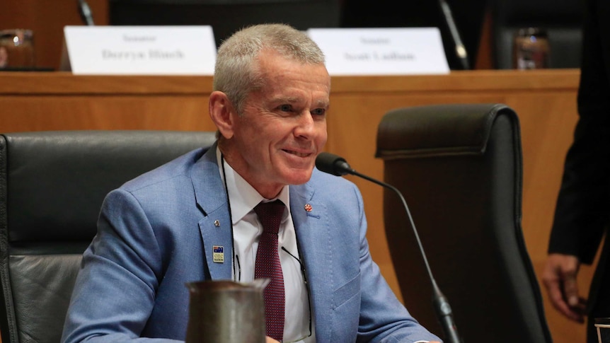 Malcolm Roberts wears a light blue suit and smiles while holding a pen in his right hand during senate estimates.