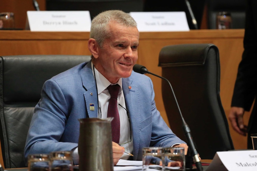 Malcolm Roberts wears a light blue suit and smiles while holding a pen in his right hand during senate estimates.