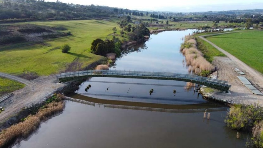 A thin bridge stretches over a brown river. Green grass grows on both sides of the river.