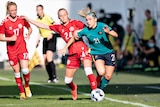A Matildas player runs with the ball with her ponytail flying behind her as a Danish defender puts in a tackle.