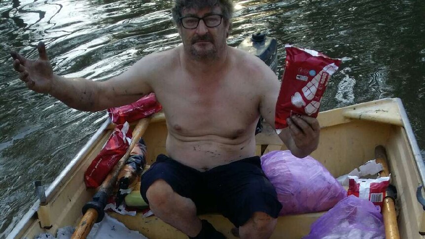 A man sits in a boat filled with garlic bread and corn on a river.