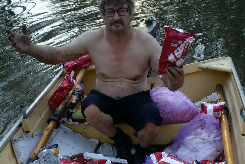 A man sits in a boat filled with garlic bread and corn on a river.
