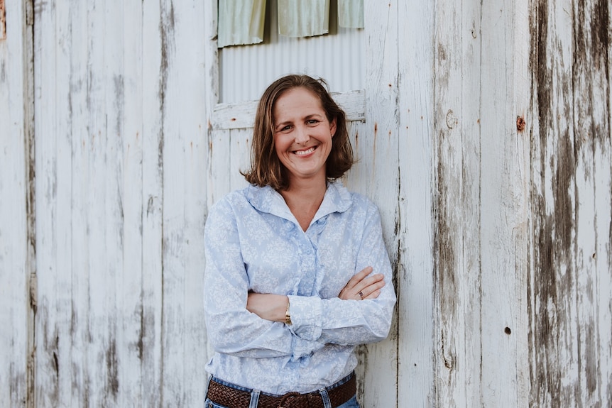 A woman in casual work shirt and jeans stands next to an old whitewashed shed.