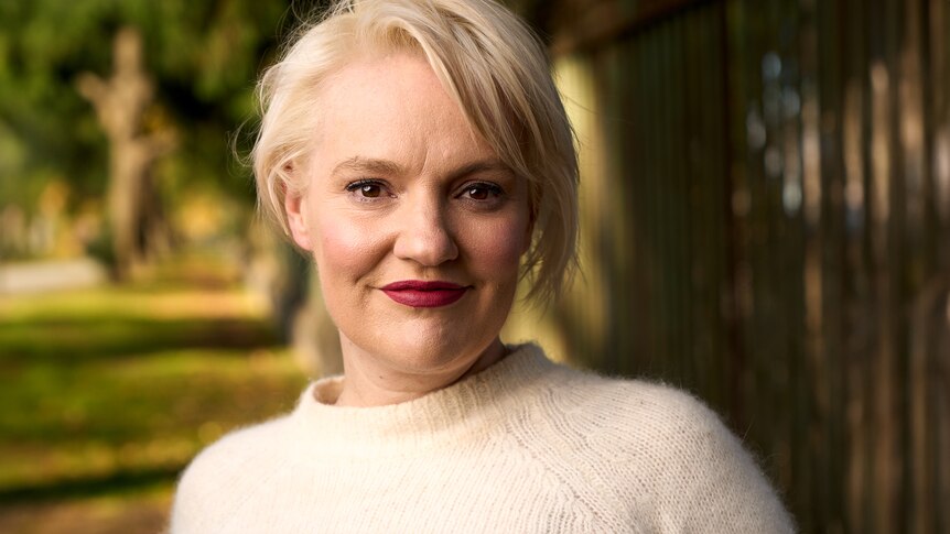 Jacinta Parsons smiling on a leafy street in a close up headshot. She wears red lipstick and a cream knitted jumper.