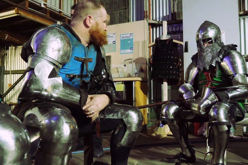Two men in suits of armour sit and talk before fighting in the ring
