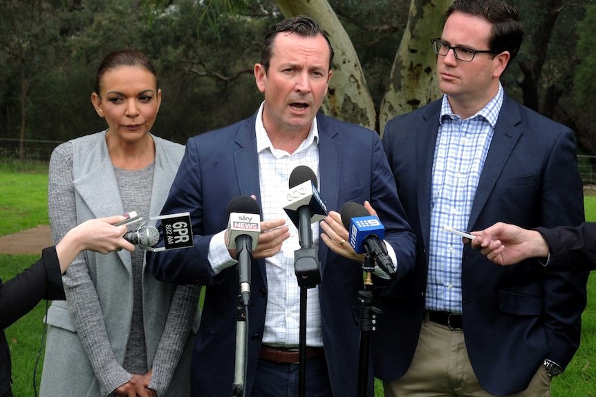 Mark McGowan stands speaking in front of a row of microphones with Anne Aly to his right and Matt Keogh to his left.