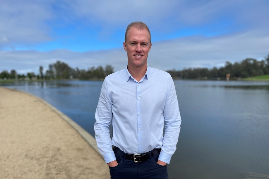 A man in a light blue shirt and black slacks stands in front of a lake