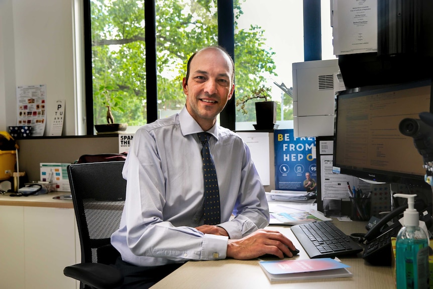 A doctor in a business shirt and tie sits in his office with computer in foreground and medical equipment in the background