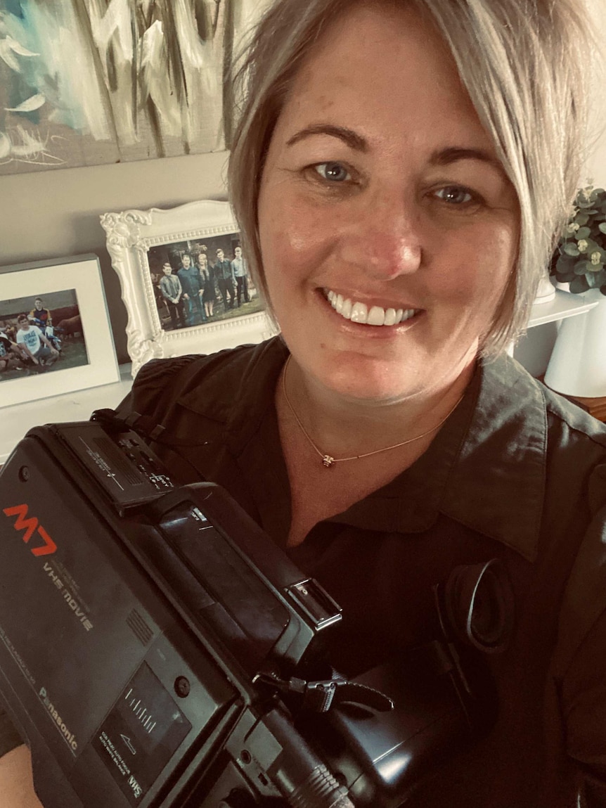 An image of a woman with short blonde hair holding an old video camera, the technology is out-of-date.