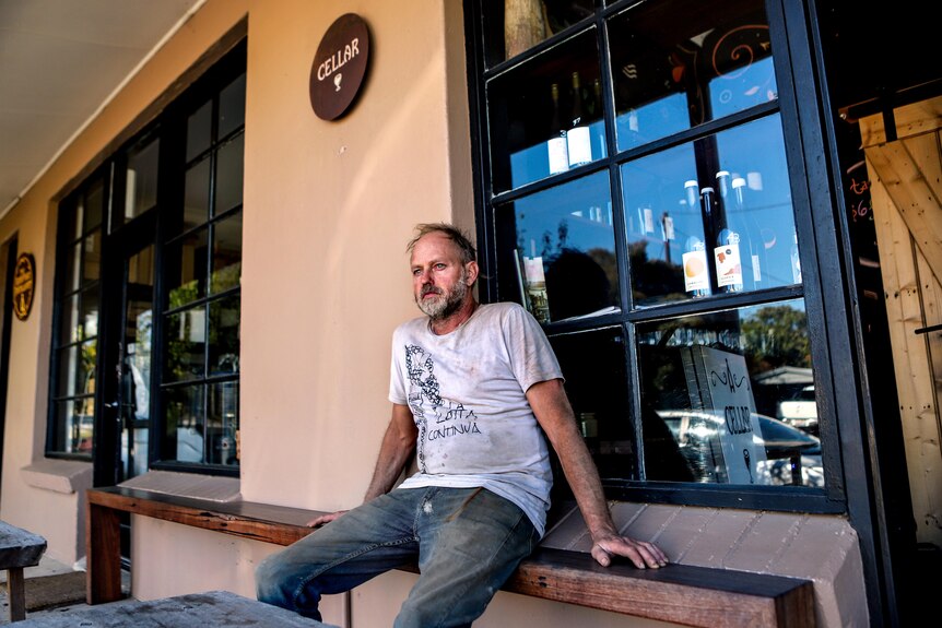 Man with thinning grey hair and white t-shirt sits outside restaurant with wine bottles in window 