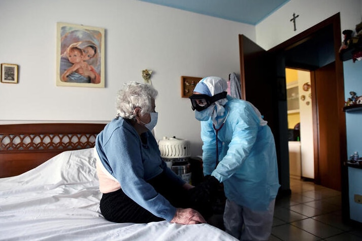 A woman in full PPE holding the hand of an elderly Italian lady sitting on her bed