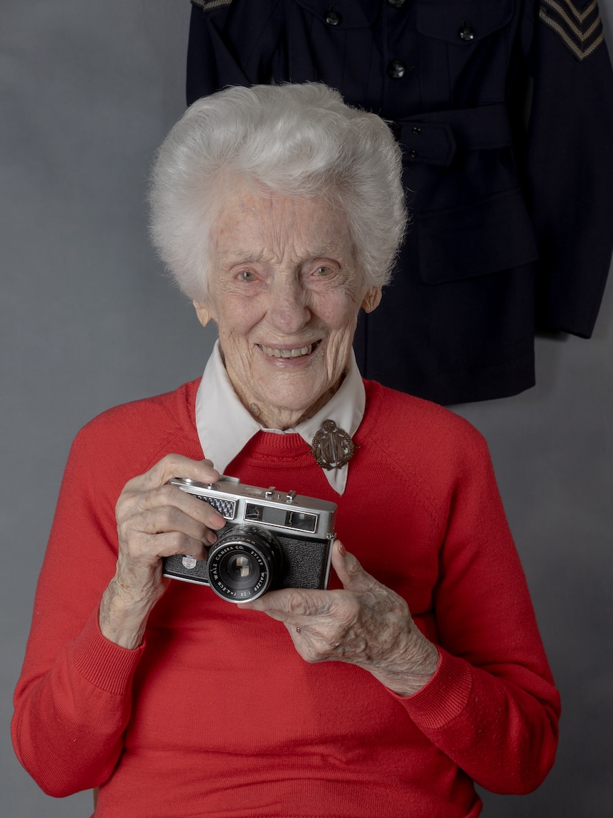 A smiling elderly woman, grey-haired bob, red sweater, cream shirt with a brooch, holds a camera.