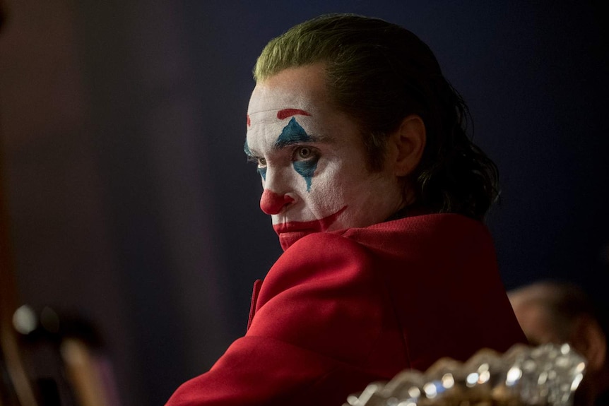 Profile of the actor in clown make-up, hair died green, sitting in chair, wearing red jacket.
