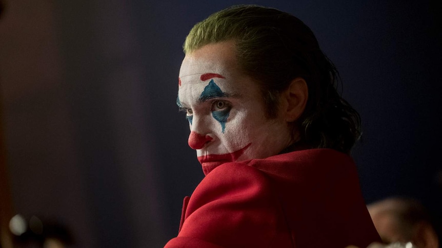 Profile of the actor Joaquin Phoenix in clown make-up, hair died green, sitting in chair, wearing red jacket.