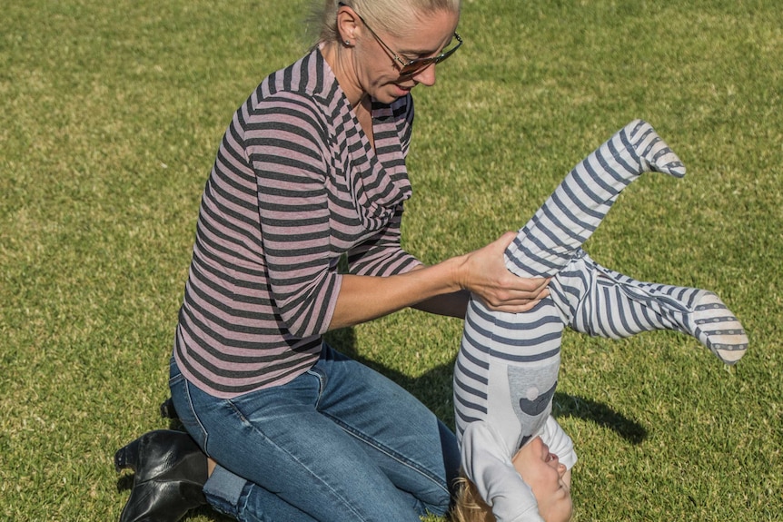 A woman in jeans and a striped shirt helps her toddler daughter do a handstand.