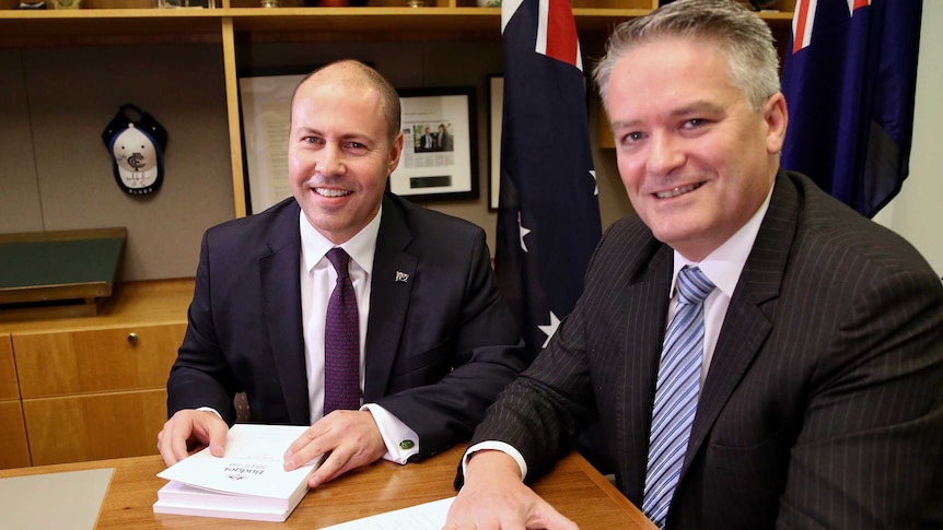 Treasurer Josh Frydenberg and Finance Minister Mathias Cormann smile at the camera while sitting at a desk with the Budget.