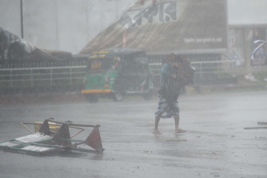 A man tried to walk across a street with umbrella in the deluge, tuk tuk in background, while a barrier has fallen over