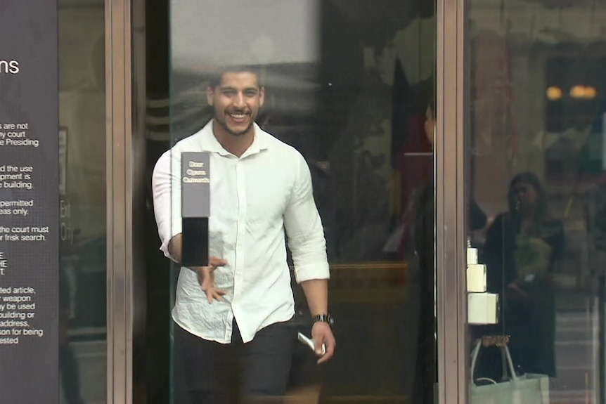 Al Zwainy wears a white shirt and black pants and smiles while opening the door of the courthouse
