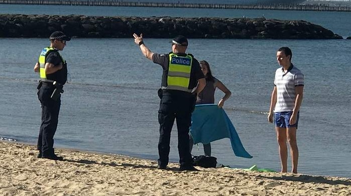Two police officers stand next a couple on the beach near the water with a cruise ship in the background.