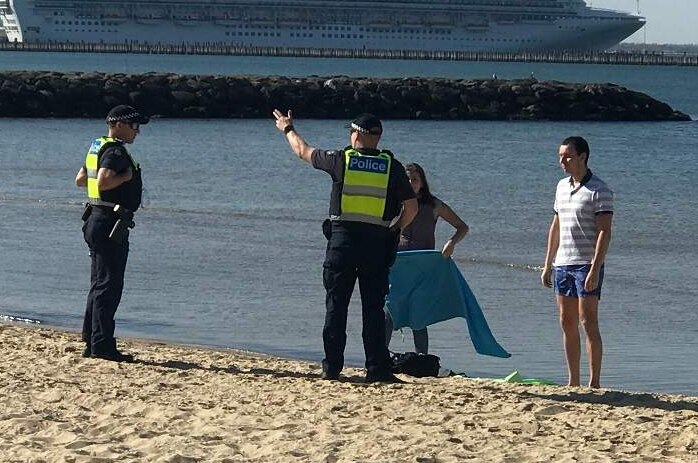 Two police officers stand next a couple on the beach near the water with a cruise ship in the background.