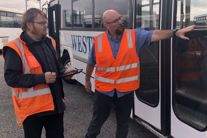 Two men wearing hi-vis vests stand inspecting a bus and looking at a glass door.